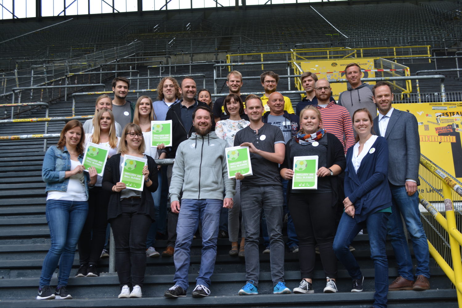 Eine Gruppe von etwa 20 Personen steht auf den Stufen eines Fußballstadions und hält jeweils ein grünes Schild mit der Aufschrift "Inklusiv am Ball bleiben" in die Kamera. Die Gruppe besteht aus Männern und Frauen unterschiedlichen Alters und mit unterschiedlicher Kleidung. Im Hintergrund sind die leeren Sitzreihen des Stadions zu sehen.