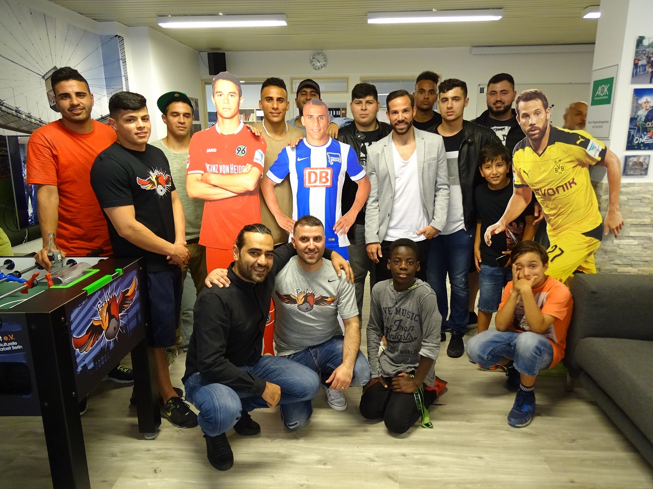 Eine Gruppe junger Männer posiert in einem Jugendtreff in Spandau. Im Hintergrund sind Poster von Fußballspielern und Fußballvereinen zu sehen. Einige tragen Fußballtrikots verschiedener Vereine.