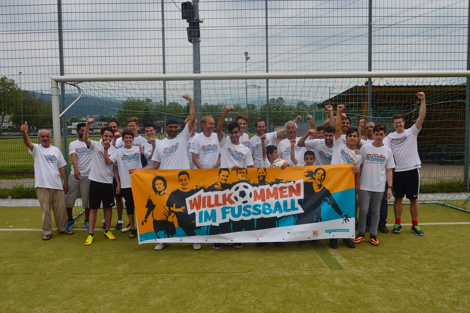 Eine Gruppe von etwa 20 Personen steht auf einem Fußballplatz vor einem Tor. Sie alle tragen weiße T-Shirts mit dem Aufdruck "Freunde statt Fremde" und lächeln in die Kamera. Vor ihnen halten sie ein großes Banner mit der Aufschrift "WILLKOMMEN IM FUSSBALL".