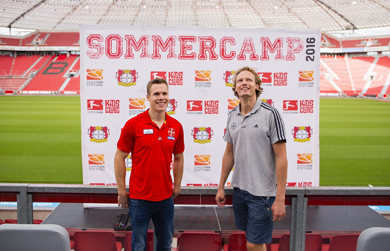 Zwei Frauen stehen auf einem Fußballfeld vor einer Werbeplane mit der Aufschrift "Kids Club Sommercamp 2016" und dem Logo der DFL Bundesligastiftung. Die Frau links trägt ein rotes Trikot mit dem Logo von Bayer Leverkusen, die Frau rechts ein graues T-Shirt. Im Hintergrund sind die leeren Zuschauerränge des Fußballstadions zu sehen.