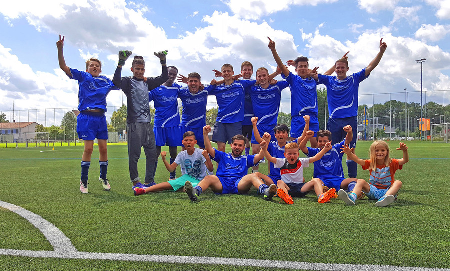 Jubelnde Kinder- und Jugendfußballmannschaft in blauen Trikots auf einem grünen Kunstrasenplatz.
