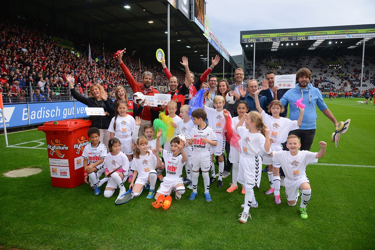 Eine Gruppe von Kindern in weißen Trikots mit dem "fit4future"-Logo steht auf dem Rasen eines Fußballstadions. Sie schauen in die Kamera und lächeln, einige heben die Hände. Im Hintergrund sind Zuschauer und das Stadion zu sehen.