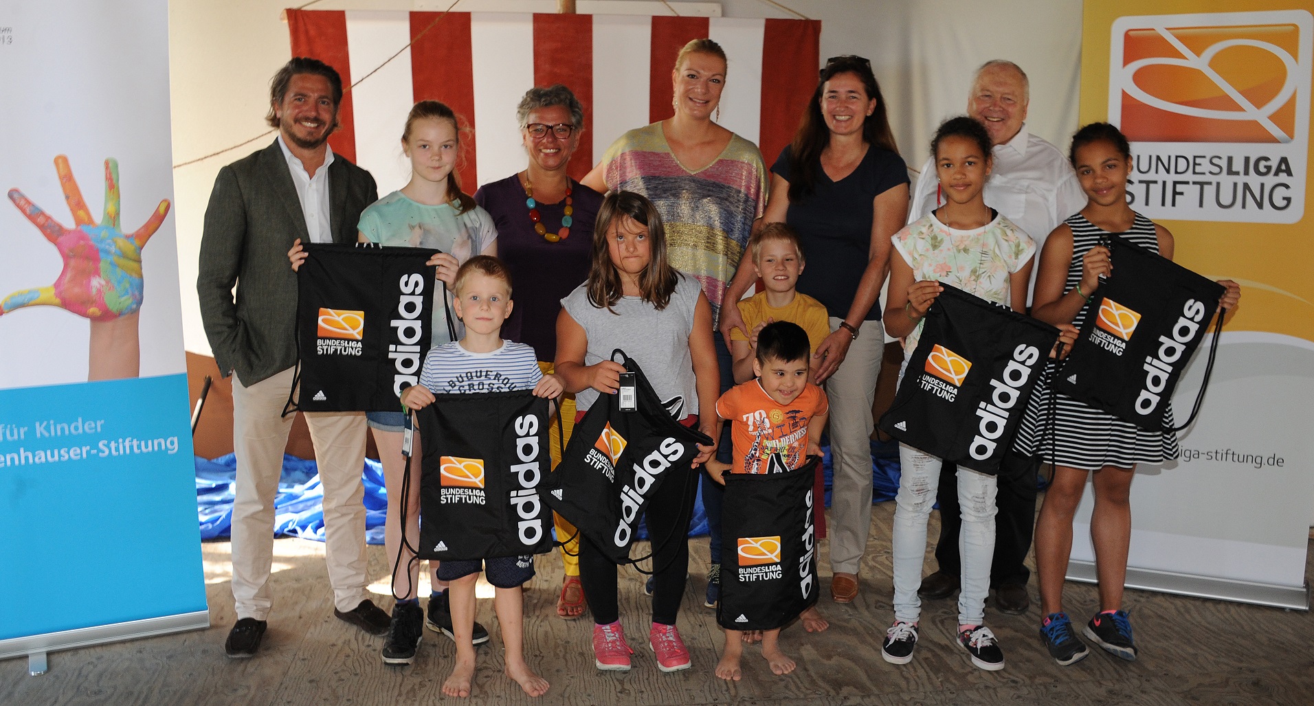 Eine Gruppe von Kindern steht lächelnd mit schwarzen Sporttaschen mit dem Logo der Bundesliga-Stiftung und adidas vor einer Werbefläche mit Handabdrücken und Logos. Maria Höfl-Riesch, eine bekannte Sportlerin, steht ebenfalls in der Gruppe.