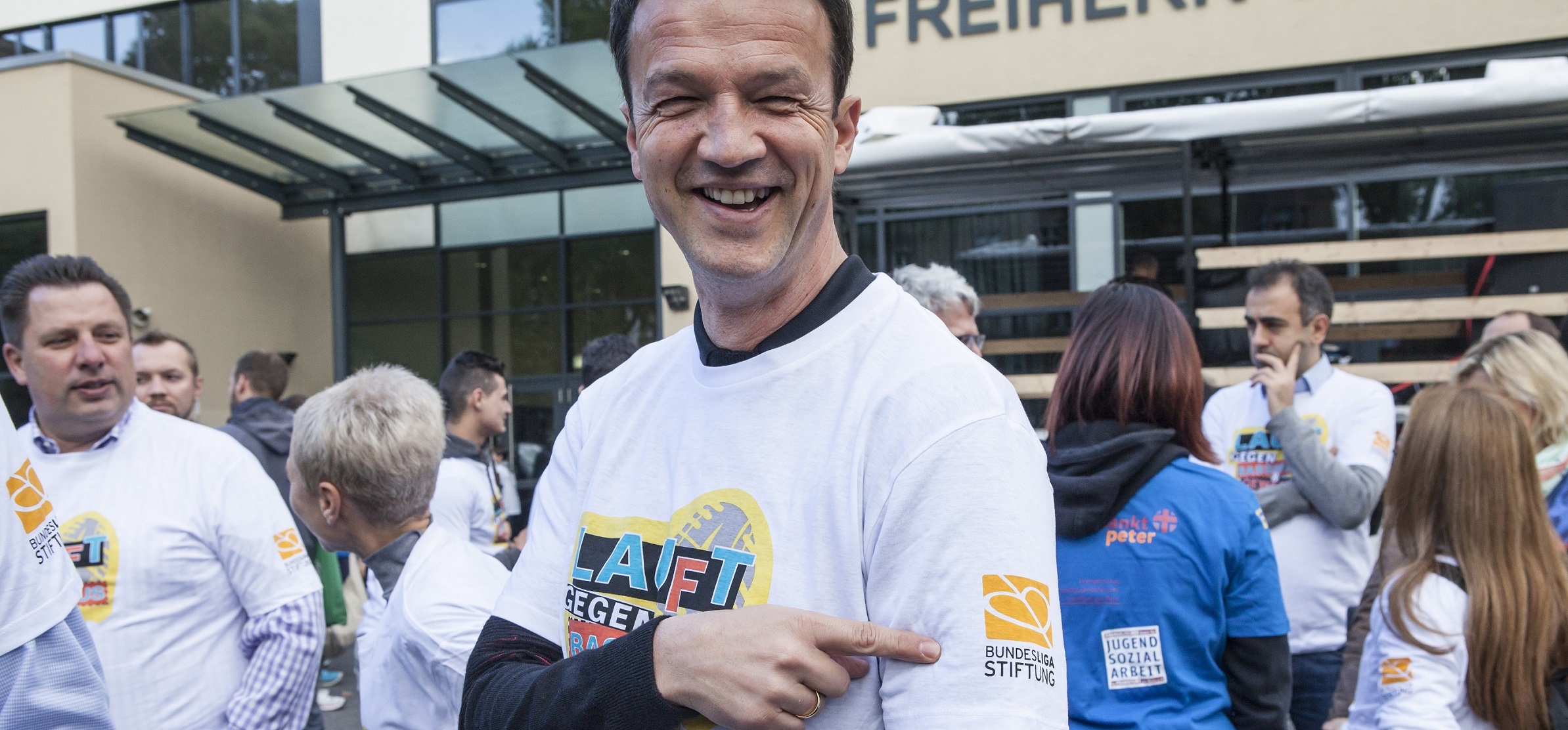Freddie Bobic nimmt an einem Lauf gegen Rassismus teil und trägt ein weißes T-Shirt mit dem Aufdruck "LAUF GEGEN" und dem Logo der DFL Stiftung. Um ihn herum laufen weitere Teilnehmer, ebenfalls in T-Shirts mit Botschaften gegen Rassismus.