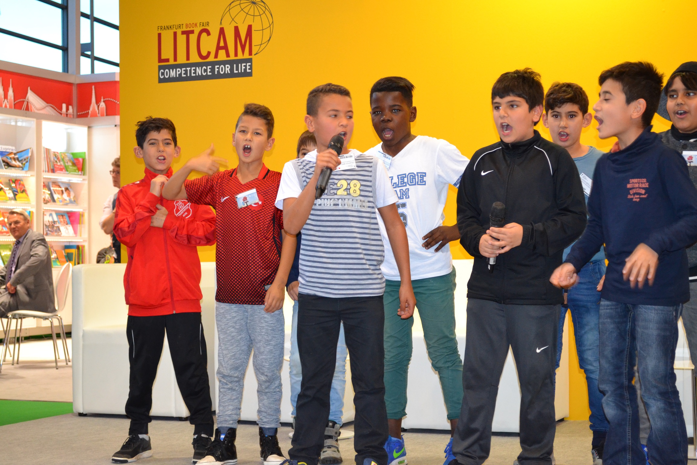 Eine Gruppe von Jugendlichen steht auf einer Bühne und hält Reden. Im Hintergrund ist ein Buchladen mit bunten Kinderbüchern zu sehen und ein Banner mit der Aufschrift "LITCAM – COMPETENCE FOR LIFE".