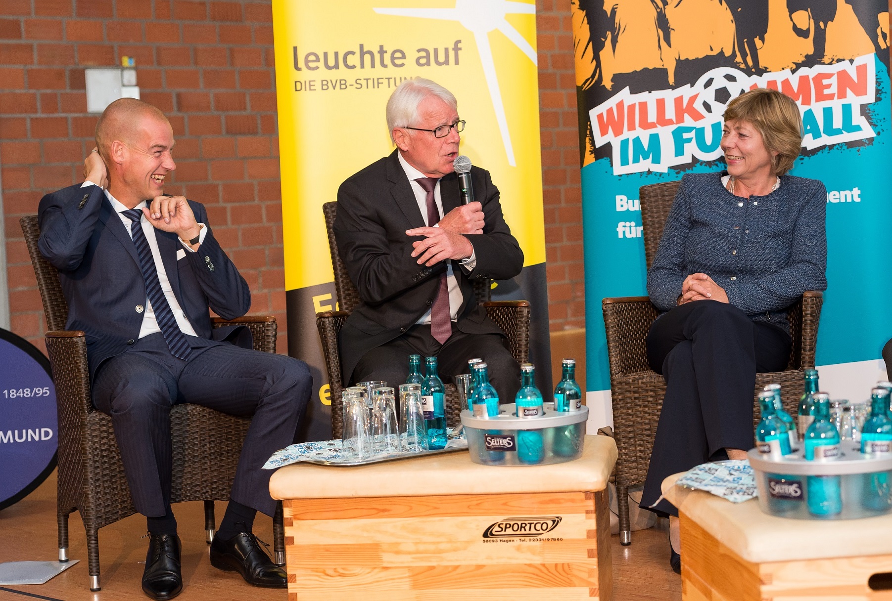 Carsten Cramer, Dr. Reinhard Rauball und Daniela Schadt sitzen nebeneinander auf einer Couch und diskutieren bei einer Veranstaltung der DFL Stiftung in Dortmund. Im Hintergrund ein Banner mit der Aufschrift "Willkommen im Fußball".