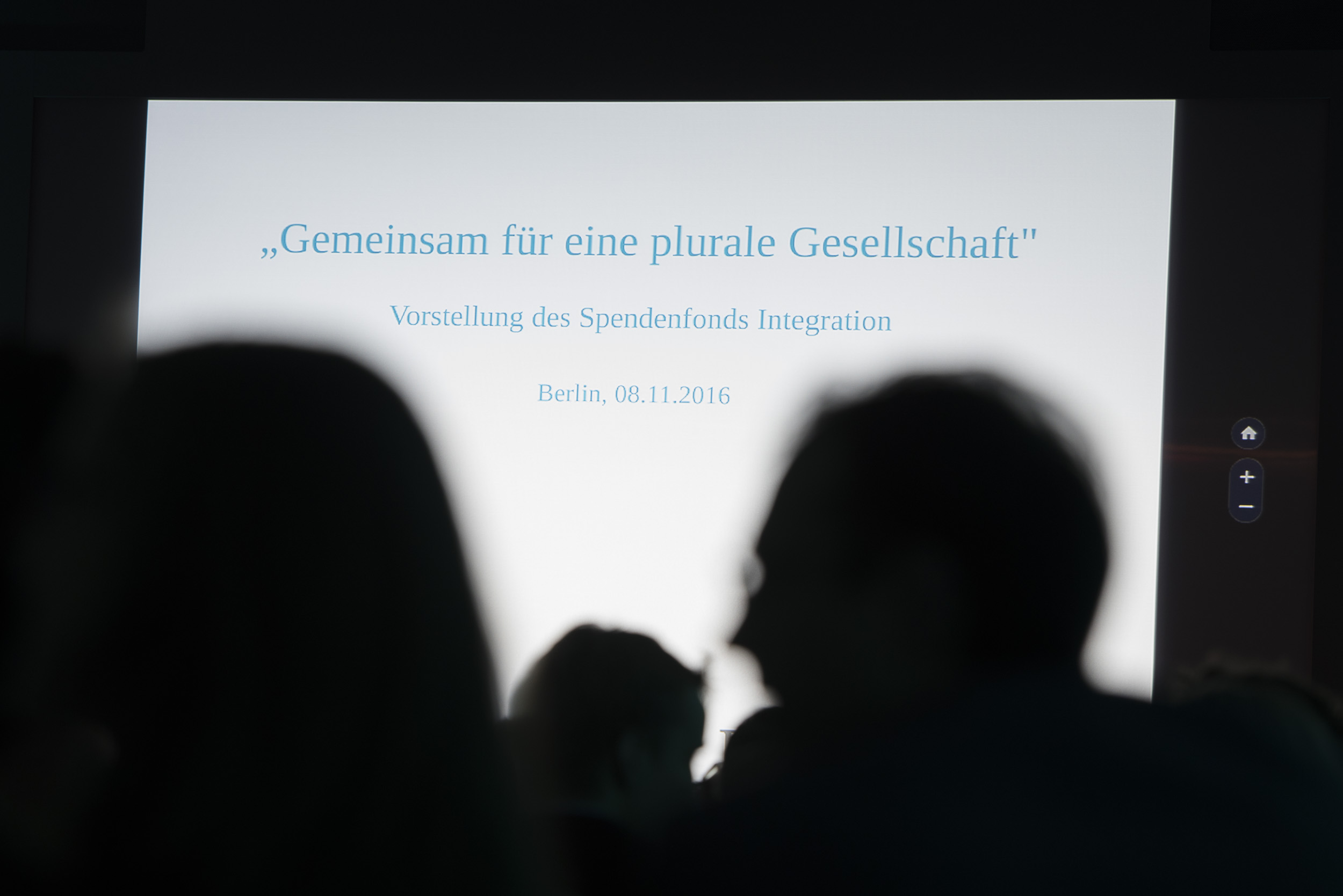 Ein dunkler Ausschnitt von zwei Personen, die einen Bildschirm betrachten, auf dem der Text "Gemeinsam für eine plurale Gesellschaft" und "Vorstellung des Spendenfonds Integration" steht. Das Bild zeigt eine Veranstaltung zur Vorstellung des Spendenfonds Integration in Berlin.