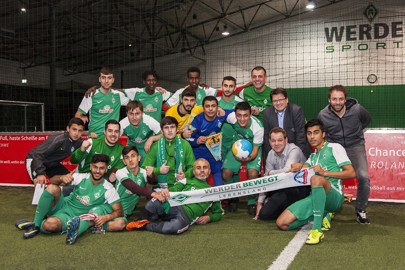 Eine Gruppe junger Männer in grünen Fußballtrikots posiert gemeinsam für ein Foto. Einige halten einen Pokal und eine Weltkugel. Im Hintergrund ist ein Banner mit der Aufschrift "WERDER BEWEGT LEBENSLANG" zu sehen.