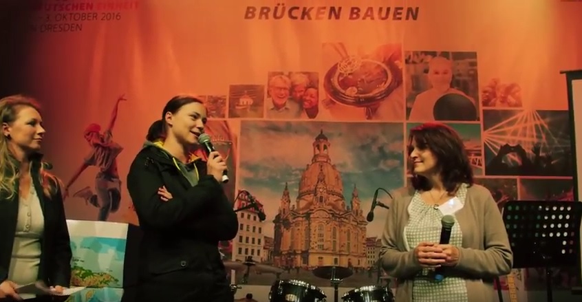 Drei Frauen stehen auf einer Bühne vor einer Kulisse mit dem Dresdner Zwinger und dem Text "Brücken bauen". Eine Frau hält ein Mikrofon und spricht, die anderen beiden stehen neben ihr und lauschen.