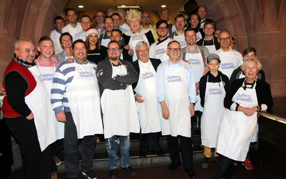 Eine große Gruppe von Menschen, viele tragen weiße Kochschürzen, posiert gemeinsam für ein Foto. Im Vordergrund stehen Männer und Frauen, die lächeln und in die Kamera schauen. Im Hintergrund sind weitere Personen zu sehen, die ebenfalls lächeln. Sie scheinen eine Veranstaltung oder ein gemeinsames Essen zu feiern.
