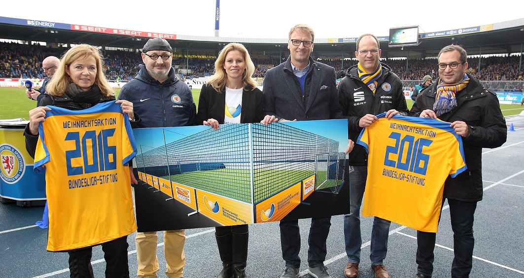 Eine Gruppe von sechs Personen hält im Stadion von Eintracht Braunschweig Trikots und eine große Pappkulisse in Form des Stadions hoch. Die Trikots und die Kulisse sind mit "Weihnachtsaktion 2016" und dem Logo der Bundesliga-Stiftung bedruckt.