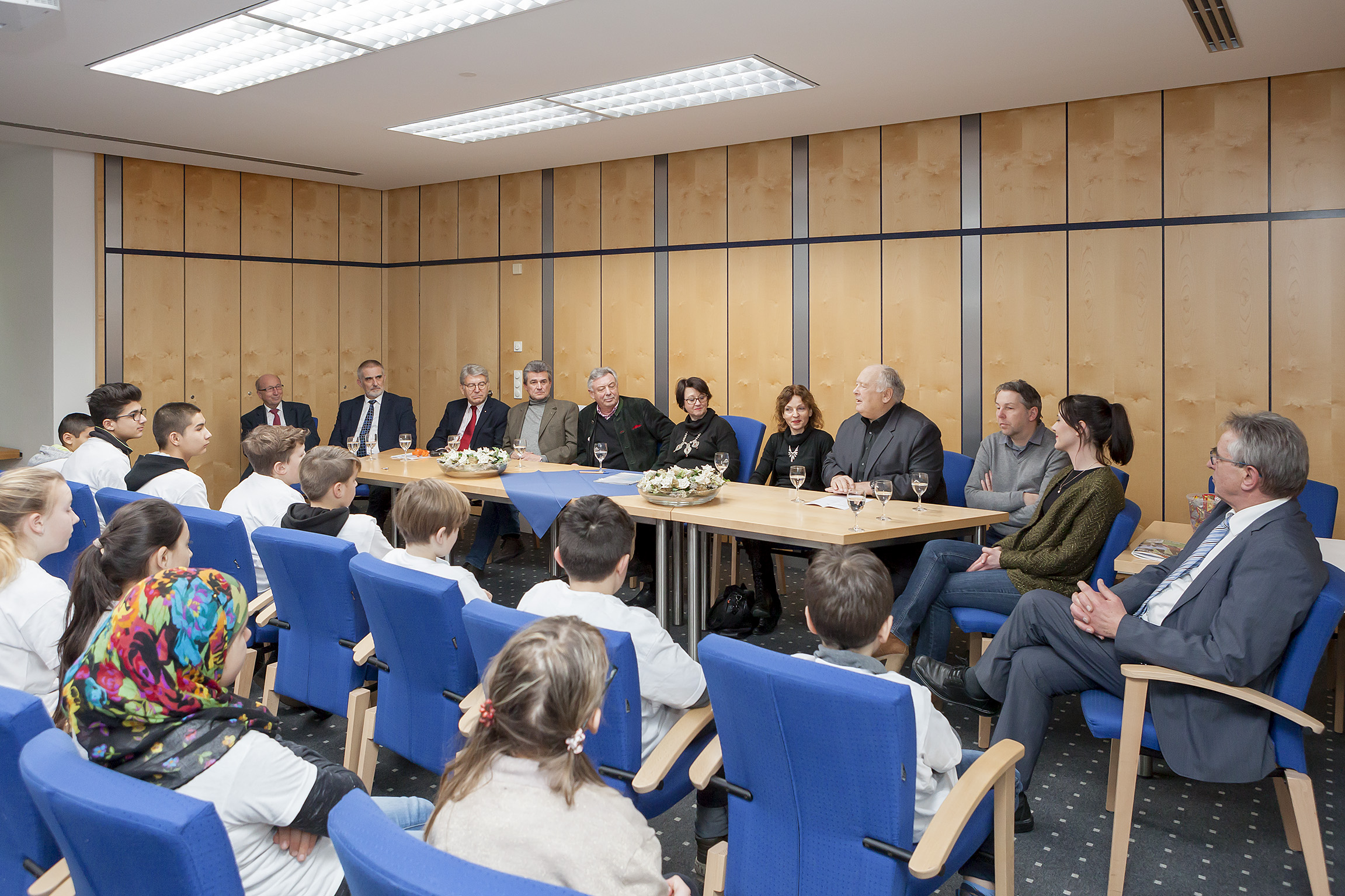 Eine Gruppe von Menschen sitzt um einen mit einer blauen Tischdecke bedeckten Tisch in einem hellen Raum. Im Vordergrund sitzen Kinder auf blauen Stühlen, während im Hintergrund Erwachsene in Anzügen und Kleidern sitzen. Die Szene zeigt eine Spendenübergabe im Rahmen der Initiative "Fußball trifft Kultur" im Rathaus Neuötting.