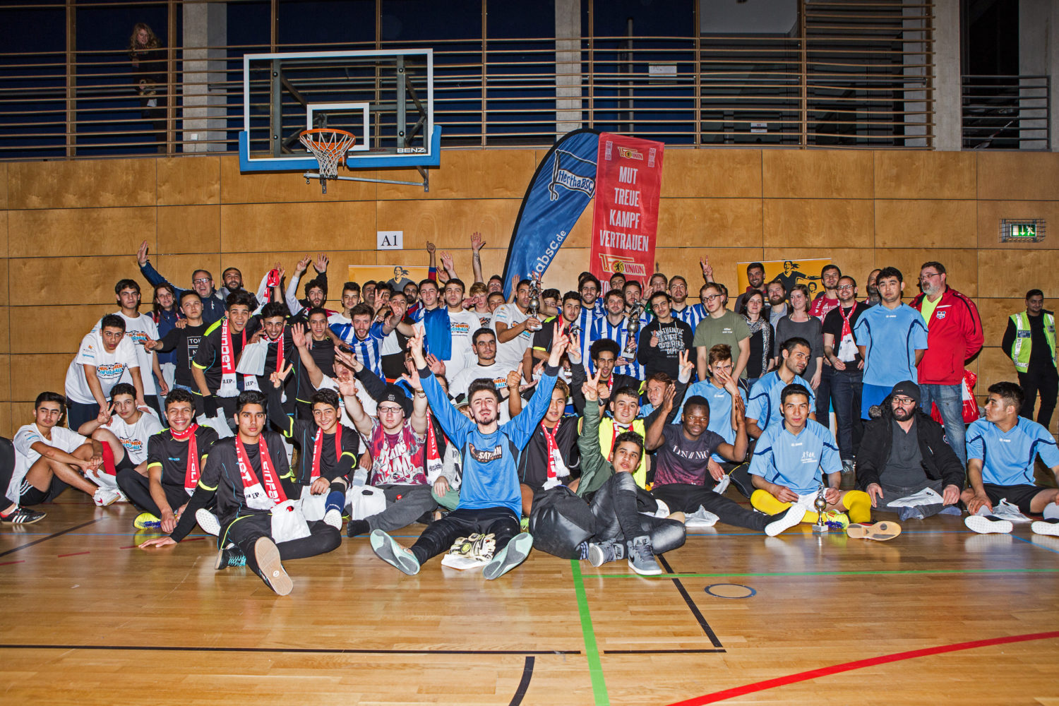 Eine große Gruppe von Jugendlichen und jungen Erwachsenen sitzt und steht in einer Turnhalle zusammen. Viele lächeln in die Kamera und zeigen Daumen hoch. Im Hintergrund ist ein Basketballkorb und ein Banner mit der Aufschrift "MUT, TREUE, KAMPF" zu sehen. Das Bild zeigt eine Aktion der DFL Stiftung mit Teilnehmern.