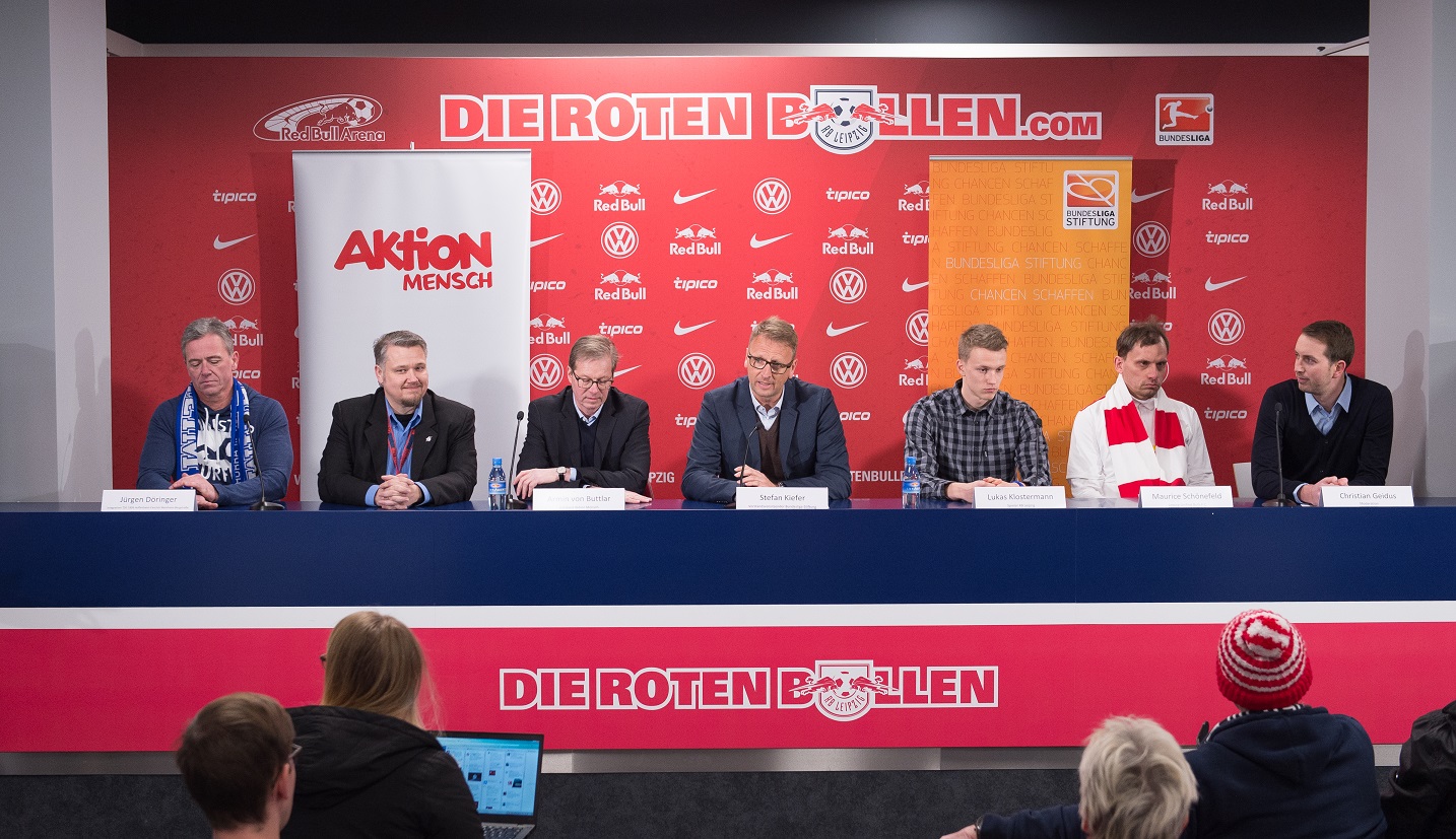 Eine Reihe von Männern sitzt an einem langen Tisch vor einer Wand mit dem Schriftzug "DIE ROTEN BULLEN". Sie nehmen an einer Besprechung teil, möglicherweise im Rahmen eines Fußballspiels der 1. Bundesliga Saison 2016/17 zwischen RB Leipzig und 1899 Hoffenheim. Im Hintergrund ist Werbung für "tipico" und das Logo der DFL Stiftung zu sehen. Das Bild dokumentiert das Projekt "Aktion Barrierefreies Stadion".