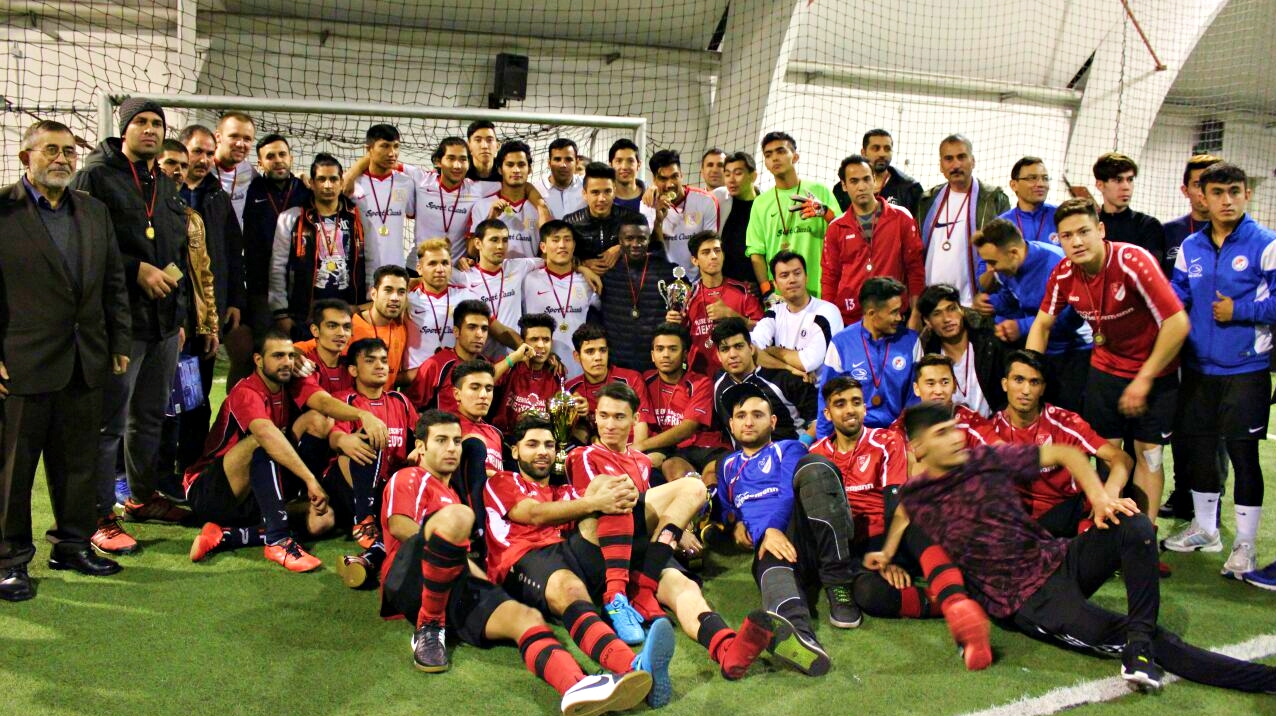 Eine große Gruppe von Fußballspielern und Betreuern posiert nach einem Turnier auf dem grünen Rasen. Viele tragen rote oder blaue Trainingsanzüge und Medaillen. Ousmann Manneh ist in der Gruppe erkennbar.