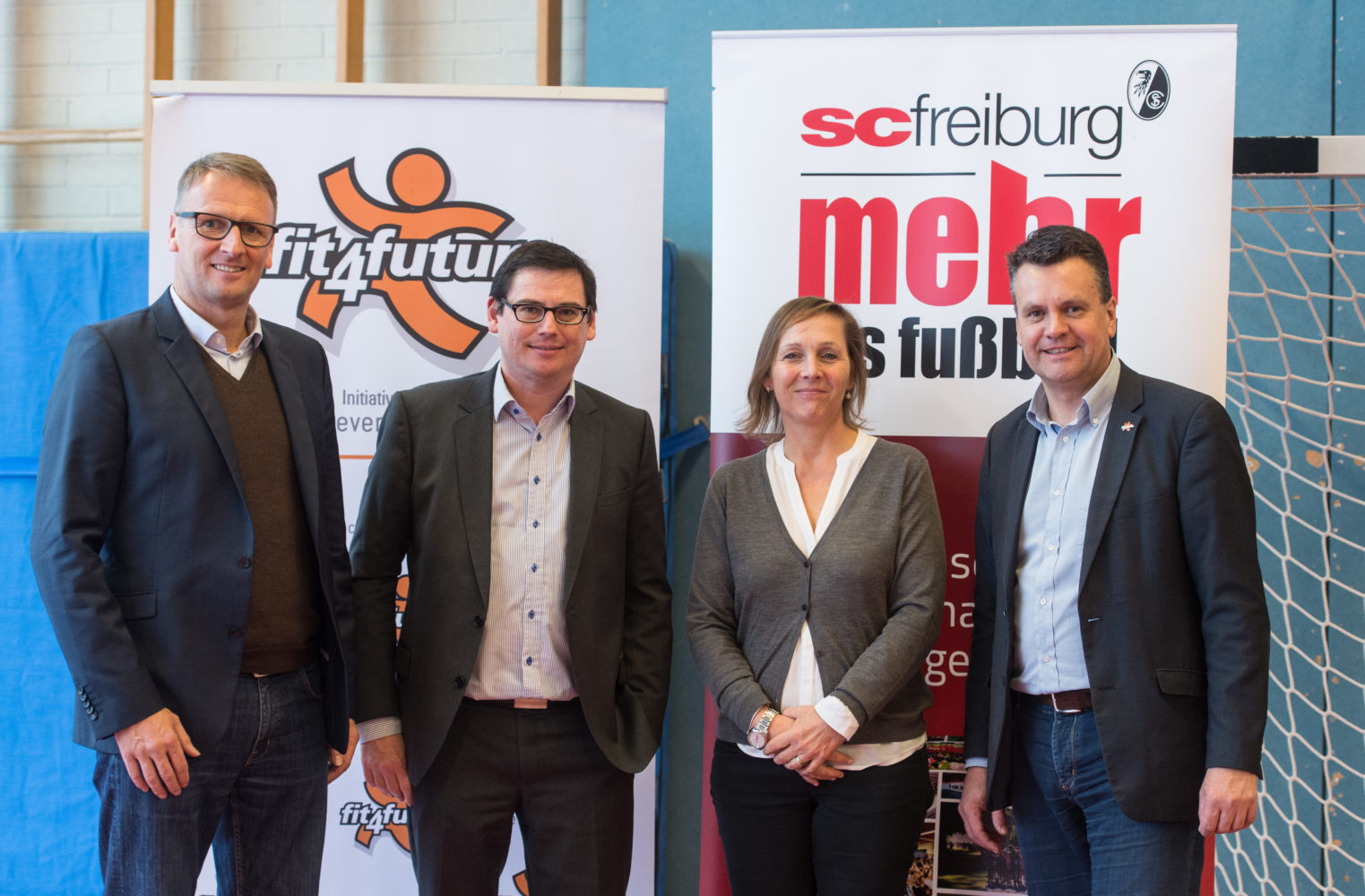 Vier Personen stehen lächelnd in einer Sporthalle. Im Hintergrund sind Banner mit den Aufschriften "fit4future" und "SC Freiburg" zu sehen. Es handelt sich um eine Veranstaltung der DFL Stiftung im Rahmen des Workshops "Bewegung" in Stegen.