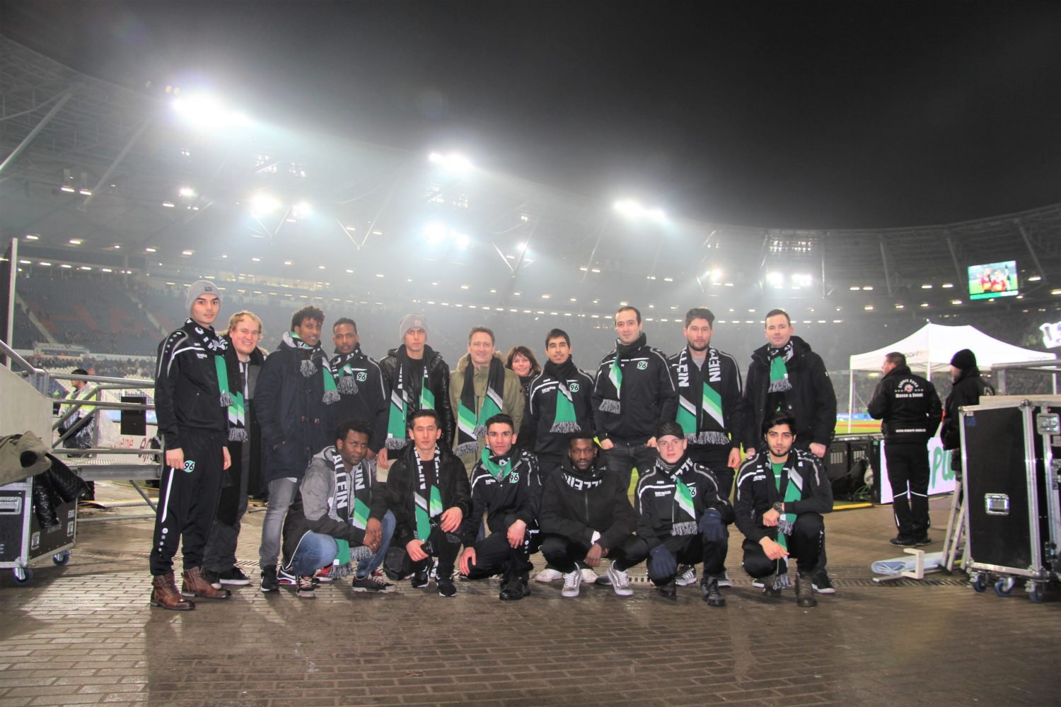 Eine Gruppe von rund 20 Personen in dunkelblauen Trainingsanzügen mit dem Aufdruck "DFL" steht und hockt auf einem Stadionvorplatz vor Scheinwerfern und Kameras. Es handelt sich wahrscheinlich um Mitarbeiter oder Helfer der DFL Stiftung bei einer Fußballveranstaltung.