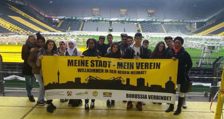 Eine Gruppe junger Menschen steht in einem Fußballstadion und hält ein Banner mit der Aufschrift "Meine Stadt - Mein Verein - Willkommen in der neuen Heimat!" und einer Skyline von Dortmund. Das Banner wird von verschiedenen Logos gesponsert.