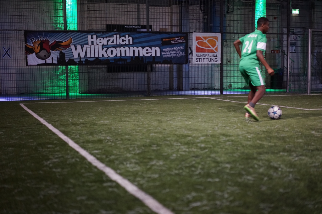 Ein junger Mann spielt Fußball auf einem beleuchteten Street-Football-Platz bei Nacht. Im Hintergrund ist ein Banner mit der Aufschrift "Herzlich Willkommen" und dem Logo der DFL-Stiftung zu sehen.