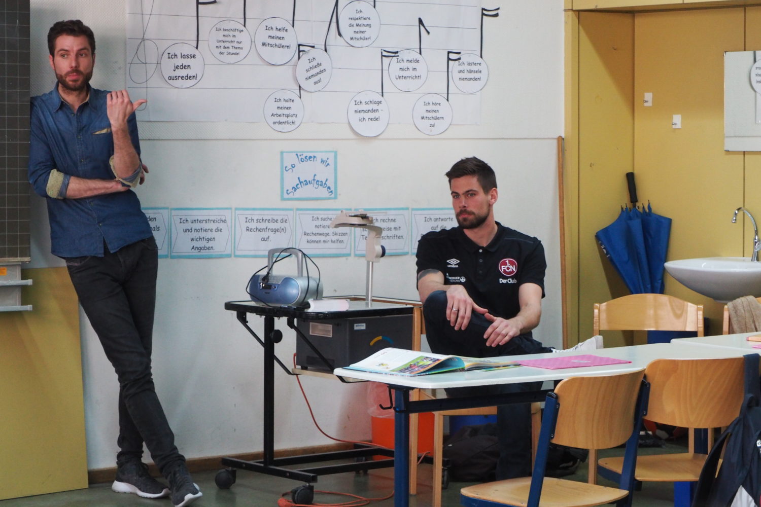 Ein Mann steht vor einer Tafel, auf der mit farbigen Stiften verschiedene Aussagen wie "Ich lasse jeden ausreden" oder "Ich schreibe auf" geschrieben stehen. Er gestikuliert mit einem Stift in der Hand und scheint etwas zu erklären. Im Hintergrund ist ein Schulklasse-Setting mit Stühlen und Tischen erkennbar, auf denen Arbeitsmaterialien liegen.
