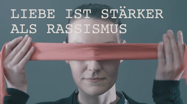Ein Mann, vermutlich Jerome Boateng, hält ein rotes Band vor sein Gesicht. Im Hintergrund steht in großen weißen Buchstaben "LIEBE IST STÄRKER ALS RASSISMUS".