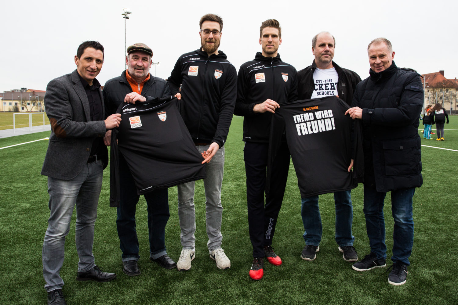 Fünf Männer stehen auf einem Fußballfeld und halten ein Trikot mit der Aufschrift "Fremd wird Freund!" hoch.