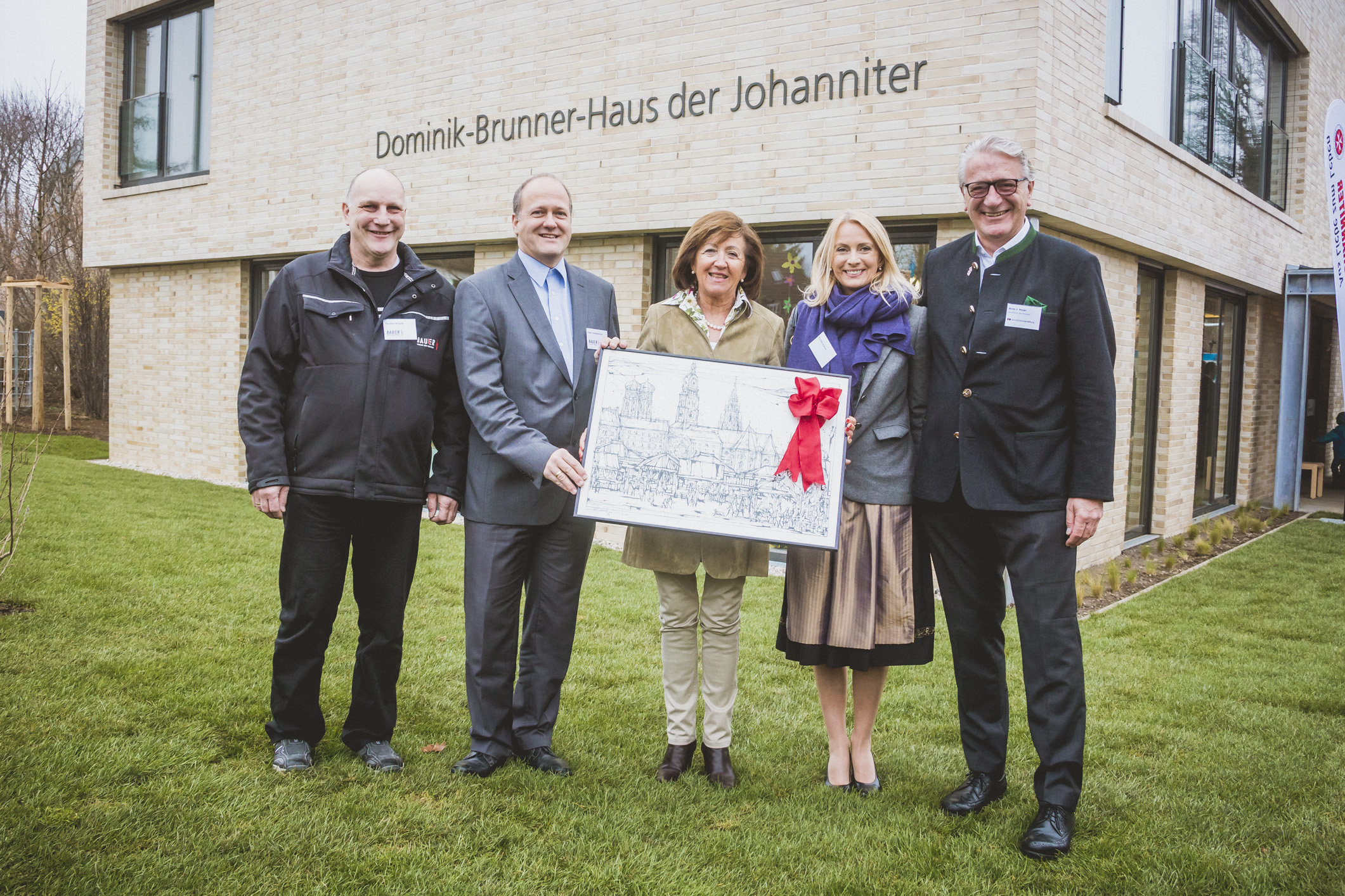 Fünf Personen stehen vor dem hellen Gebäude des "Dominik-Brunner-Hauses" der Johanniter. In der Mitte halten zwei Frauen eine gerahmte Zeichnung oder einen Plan des Gebäudes. Die anderen drei Personen stehen seitlich und lächeln in die Kamera.
