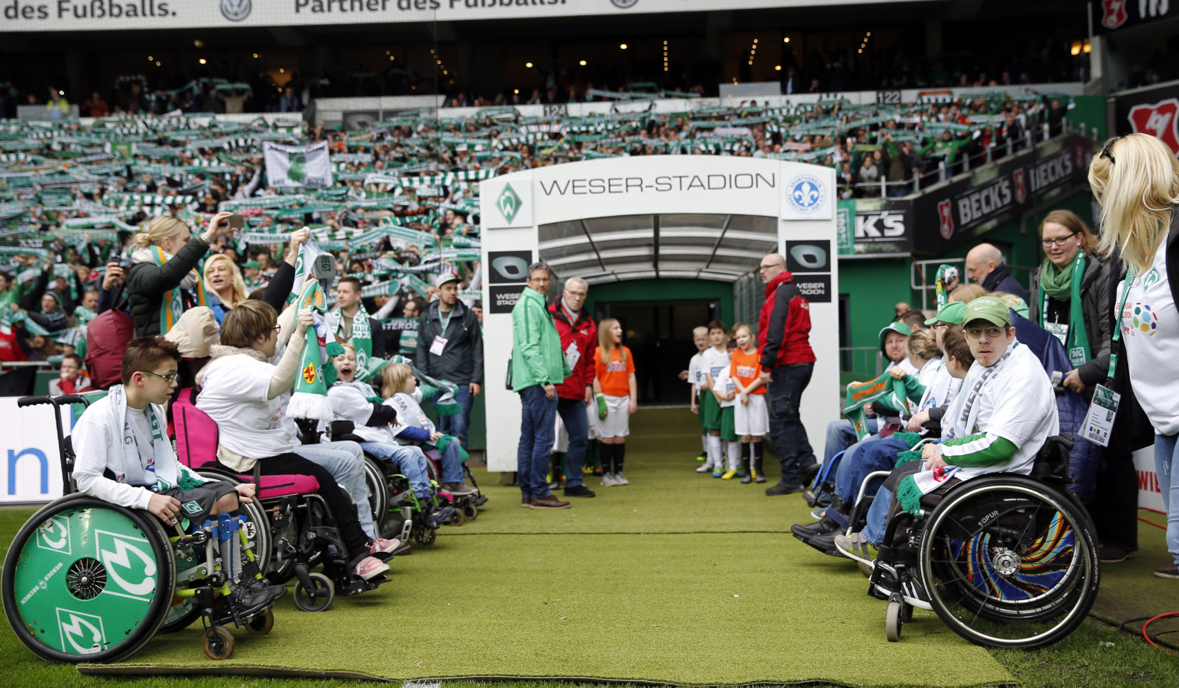 Inklusion beim Heimspiel von Werder Bremen: Rollstuhlfahrer*innen werden im Weserstadion auf dem Rasen begrüßt, umgeben von jubelnden Fans und dem Werder-Logo.