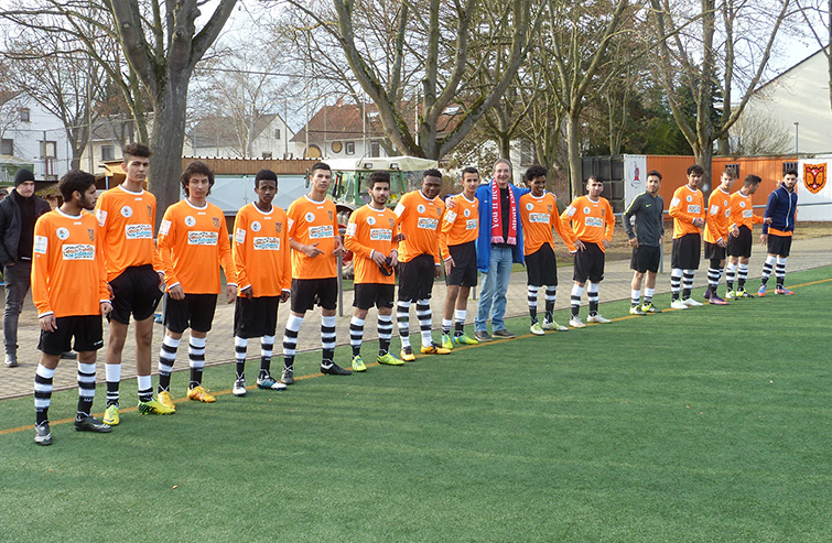 Eine Gruppe Jugendlicher in Fußballtrikots steht auf einem grünen Kunstrasenplatz aufgereiht und blickt in eine Richtung. Die Trikots sind orange mit schwarzen Akzenten. Im Hintergrund ist eine Sportanlage und teilweise Baumbestand zu erkennen.