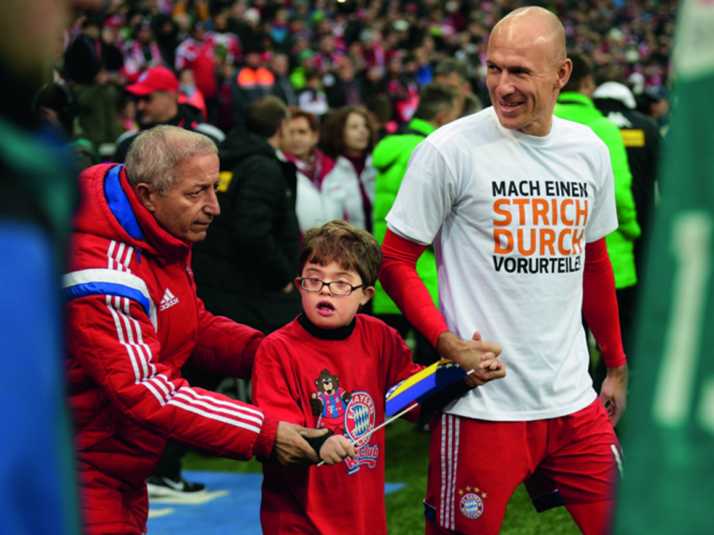 Arjen Robben führt einen Jungen mit Down-Syndrom an der Hand auf das Spielfeld. Beide tragen rote T-Shirts mit dem Logo des FC Bayern München. Robben trägt zusätzlich ein T-Shirt mit der Aufschrift "Mach einen Strich durch Vorurteile". Im Hintergrund sind weitere Personen und das Stadion zu sehen.