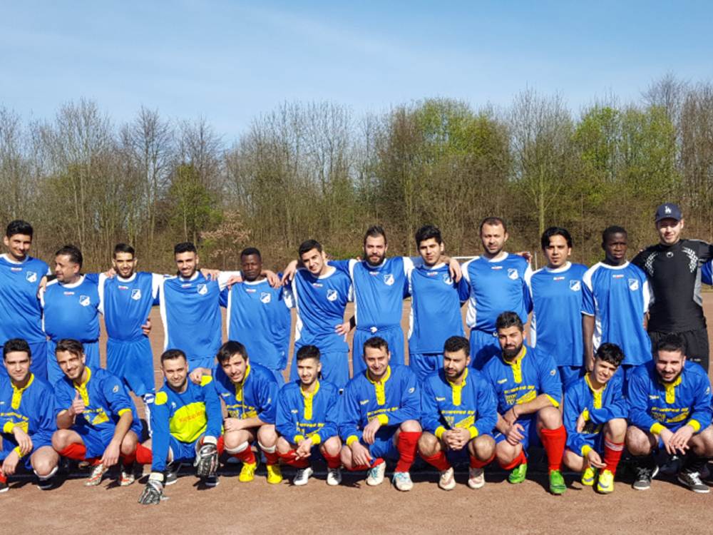 Mannschaftsfoto einer Fußballgruppe in blauen Trikots und roten Socken auf einem sandfarbenen Feld. Einige Spieler hocken, andere stehen und lächeln in die Kamera. Im Hintergrund ist eine Baumreihe zu sehen.