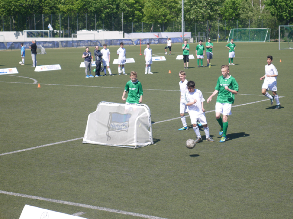 Eine Gruppe Jugendlicher spielt Fußball auf einem grünen Rasenplatz. Einige Spieler tragen grüne Trikots, andere weiße. Im Hintergrund sind weitere Personen und ein Fußballtor zu sehen.