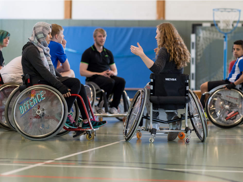 Eine Frau mit roten Locken spricht in einer Sporthalle. Sie sitzt im Rollstuhl und gestikuliert, während sie sich einer Gruppe von Menschen zuwendet, die ebenfalls im Rollstuhl sitzen. Im Hintergrund sind weitere Personen und ein Basketballkorb zu sehen. Das Bild zeigt eine inklusive Sportveranstaltung oder ein Training.