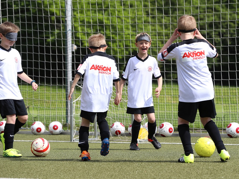 Vier Kinder in weißen und schwarzen Fußballtrikots stehen auf einem grünen Kunstrasenplatz und schauen in unterschiedliche Richtungen. Einige der Kinder schauen erwartungsvoll, während eines die Hände vor die Augen hält. Im Hintergrund steht ein Fußballtor und mehrere Fußbälle liegen auf dem Rasen.