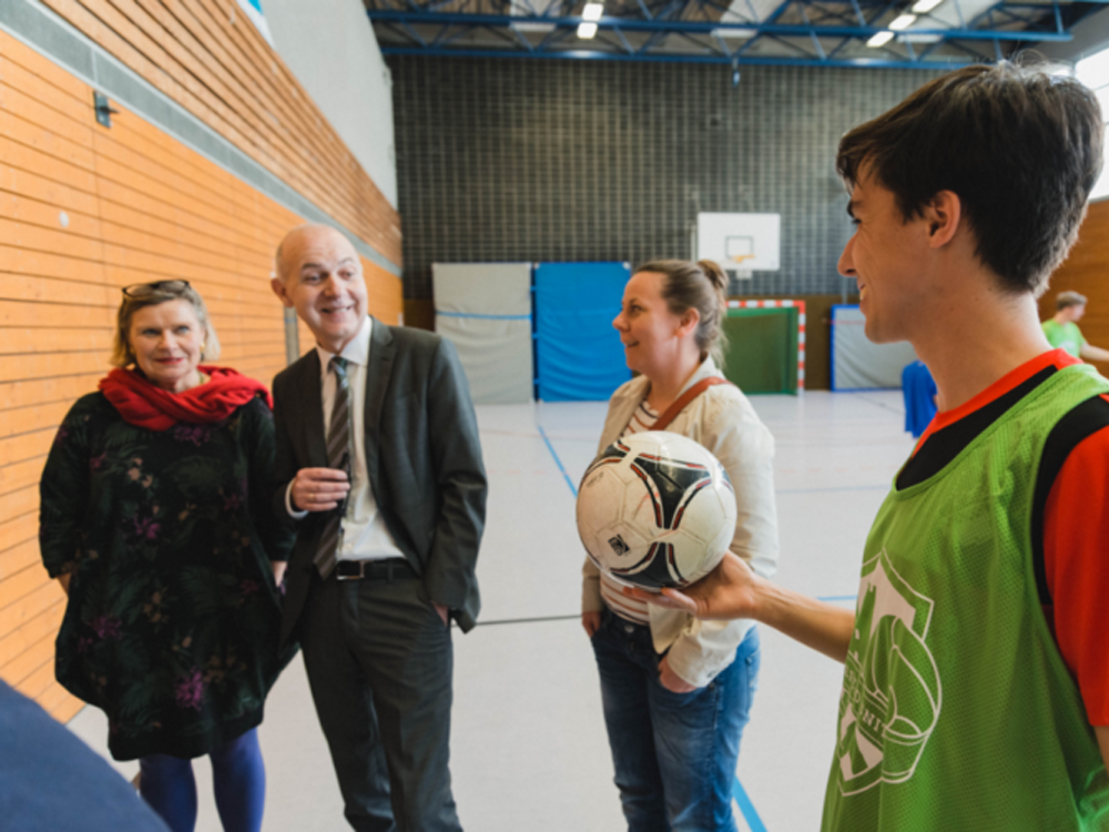 Eine Frau mit rotem Schal, ein Mann im Anzug und eine Frau mit einem Kind im Arm stehen in einer Sporthalle und unterhalten sich. Ein Jugendlicher in einer grünen Trainingsweste hält einen Fußball entgegen.