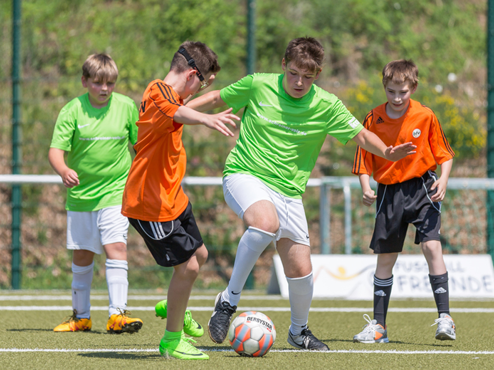 Vier Jugendliche spielen Fußball auf einem grünen Rasenplatz. Zwei Spieler in orangefarbenen und grünen Trikots kämpfen um den Ball, während die anderen beiden als Zuschauer im Hintergrund stehen.