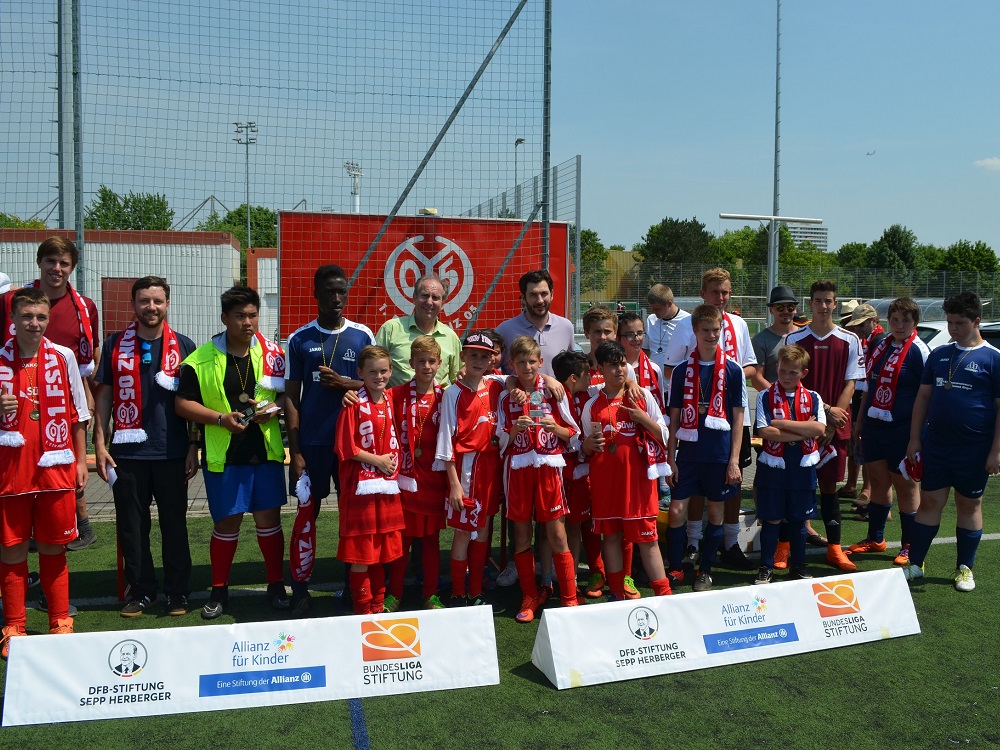 Eine Gruppe Jugendfußballspieler in roten und blauen Trikots posiert mit Sandro Schwarz, Trainer des 1. FSV Mainz 05, auf einem Fußballfeld. Im Hintergrund sind Werbebanner der DFL-Stiftung und Allianz für Kinder zu sehen.