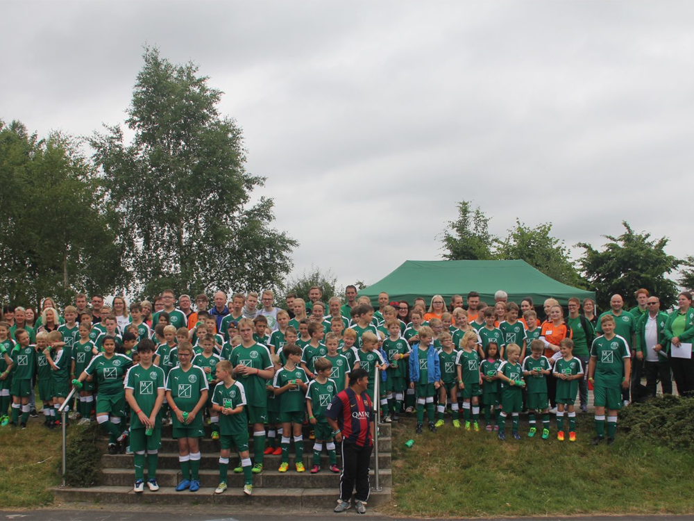 Eine große Gruppe von Kindern und Jugendlichen in Fußballtrikots steht vor einem Zelt und einer Grünfläche. Viele Erwachsene stehen ebenfalls in der Gruppe.