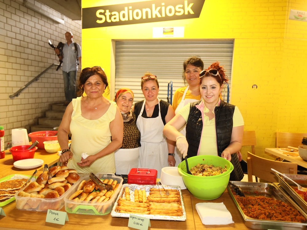 Freiwillige Frauen stehen hinter einem Imbissstand mit verschiedenen Speisen wie Brot, Gebäck und Salaten. Im Hintergrund ist ein Stadion zu erkennen.