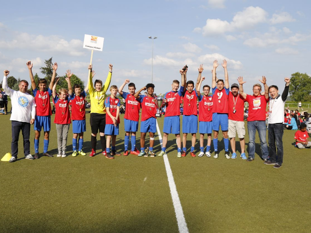 Eine Gruppe Jugendlicher in Fußballtrikots steht auf einem grünen Kunstrasenplatz und hebt die Arme in die Höhe. Einige tragen Medaillen um den Hals. Im Hintergrund sind Zuschauer und Bäume zu sehen.