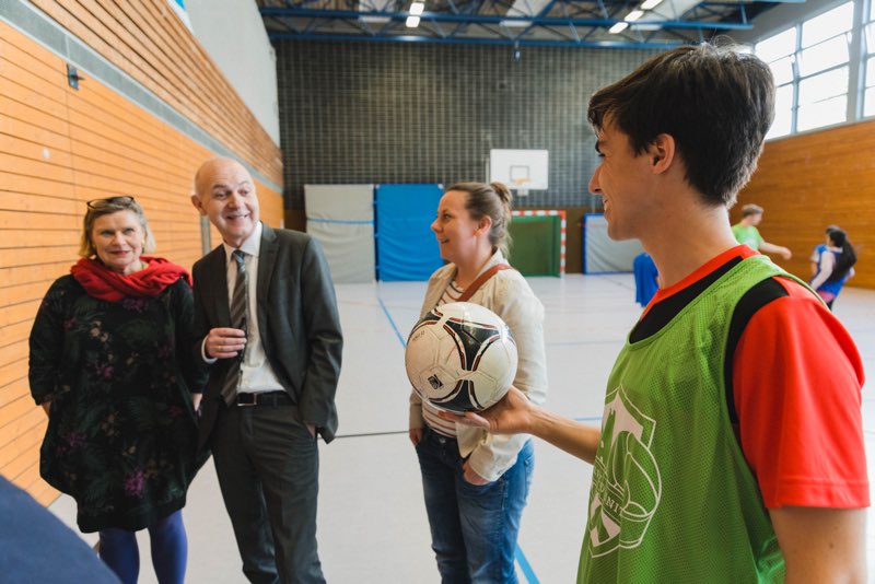 Eine Gruppe von Menschen steht in einer Sporthalle zusammen. Ein Mann im Anzug spricht mit einer Frau, während ein Jugendlicher im roten und grünen Trainingsshirt einen Fußball hält. Im Hintergrund sind weitere Personen und ein Basketballkorb zu sehen.