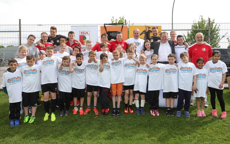 Eine Gruppe von Kindern steht auf einem Fußballplatz vor einer Bande. Viele der Kinder tragen T-Shirts mit dem Aufdruck "In safe hands". Im Hintergrund stehen Erwachsene, darunter vermutlich Fußballspieler und Betreuer. Im Hintergrund sind Werbebanner zu sehen.
