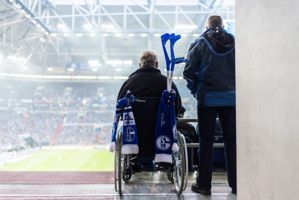 Eine Person im Rollstuhl wird von einer weiteren Person begleitet, während sie auf das Fußballfeld im Veltins-Arena Stadion in Gelsenkirchen blickt.