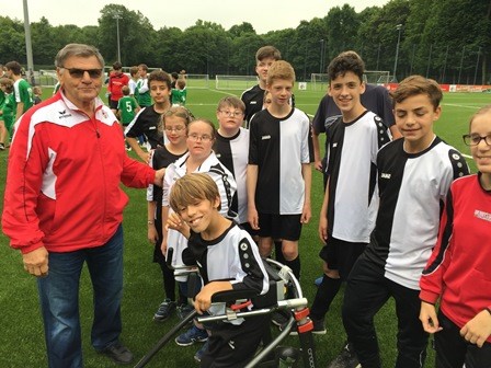 Eine Gruppe Jugendlicher, darunter ein Kind im Rollstuhl, steht auf einem Fußballfeld mit Wolfgang Weber, einem Mann in roter Trainingsjacke. Sie lächeln in die Kamera. Im Hintergrund sind weitere Personen und das Spielfeld zu sehen.