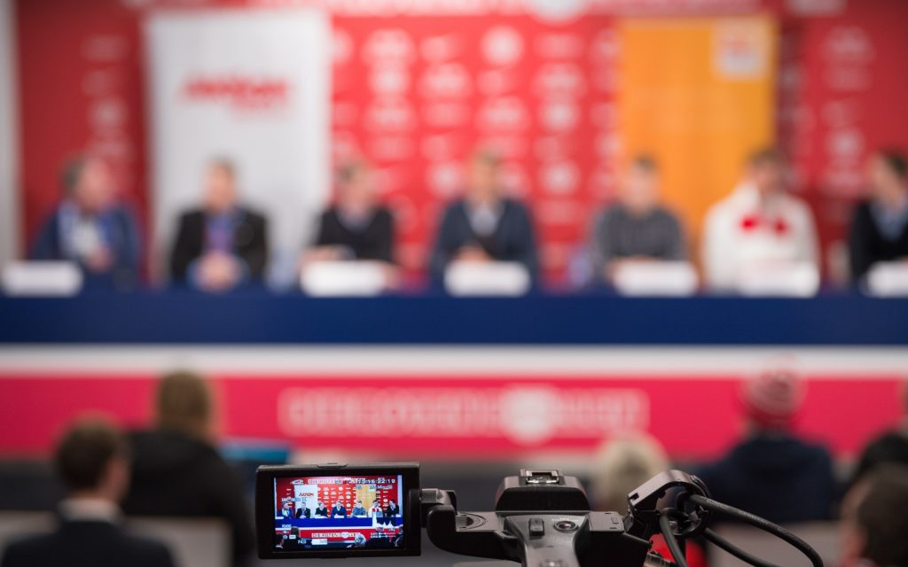 Eine Reihe von Personen sitzt an einem Tisch vor rotem Hintergrund, vermutlich bei einer Pressekonferenz. Im Vordergrund ist ein Kameramann mit seinem Equipment zu sehen, dessen Monitor das Bild der Sitzung zeigt.