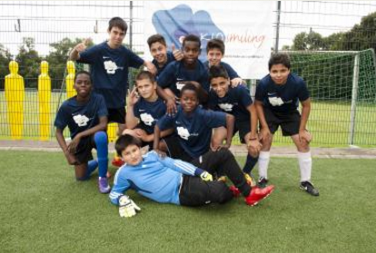 Eine Gruppe von Jugendlichen im Fußballtrikot posiert auf einem grünen Kunstrasenplatz. Ein Junge im Torwarttrikot liegt im Vordergrund, die anderen stehen oder hocken um ihn herum und lächeln in die Kamera. Im Hintergrund sind gelbe Markierungshütchen und ein Fußballtor zu sehen.