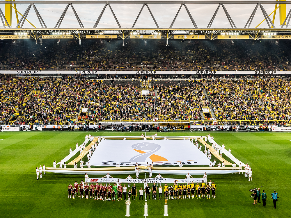 Fußballstadion gefüllt mit Zuschauern, auf dem Rasen eine große Banneraufschrift "SUPER CUP" mit den Mannschaften und offiziellen Personen gruppiert davor.