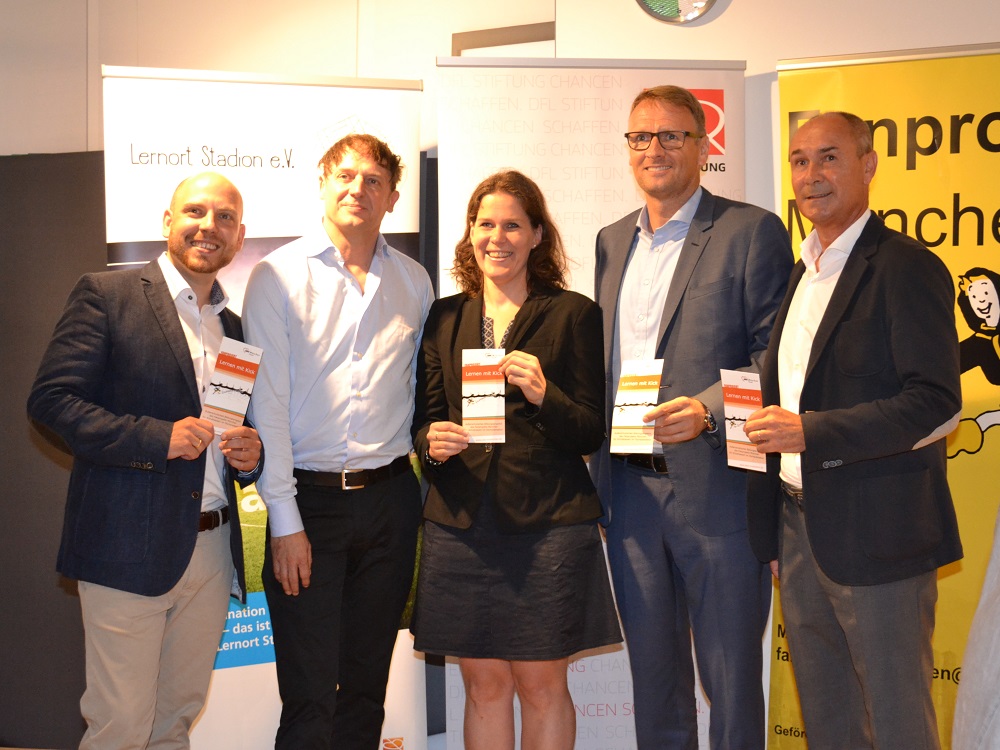 Fünf Personen stehen lächelnd vor einer Wand mit Werbeplakaten. Alle halten jeweils einen kleinen, rechteckigen Gegenstand in den Händen.