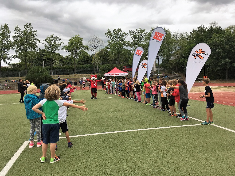 Eine Gruppe von Kindern steht auf einem grünen Sportplatz im Kreis und streckt die Arme aus. Im Hintergrund sind weitere Kinder und Betreuer zu sehen sowie mehrere Banner mit dem Logo "fit4future".