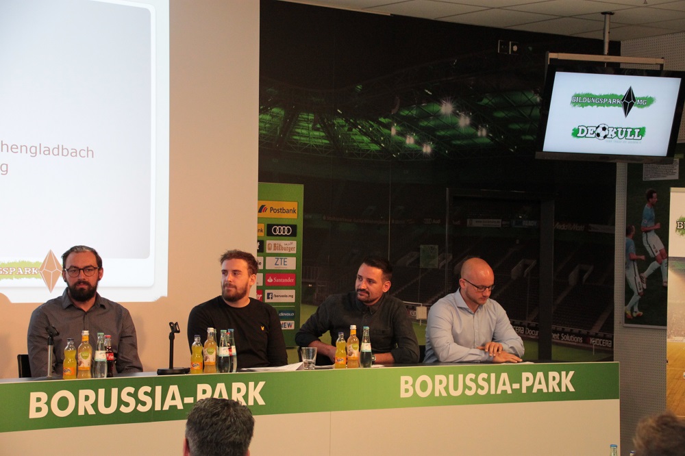 Eine Podiumsdiskussion im Borussia-Park Stadion. Vier Männer sitzen an einem Tisch mit dem Schriftzug "BORUSSIA-PARK" vor einer Wand mit Sponsorlogos und einem Bildschirm, der eine Fußballszene zeigt.